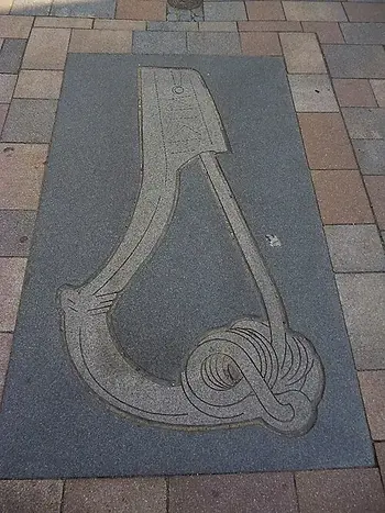 Steinerne Reliefplatte im Pflaster mit abstraktem, schlingenförmigem Motiv in Grau und Beige.